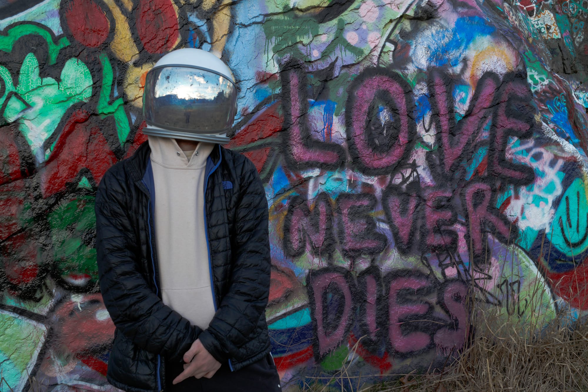A person in a white vintage space helmet with a mirrored visor and a black puffer jacket, standing against a rock wall covered in graffiti, with LOVE NEVER DIES sprayed in pink beside them