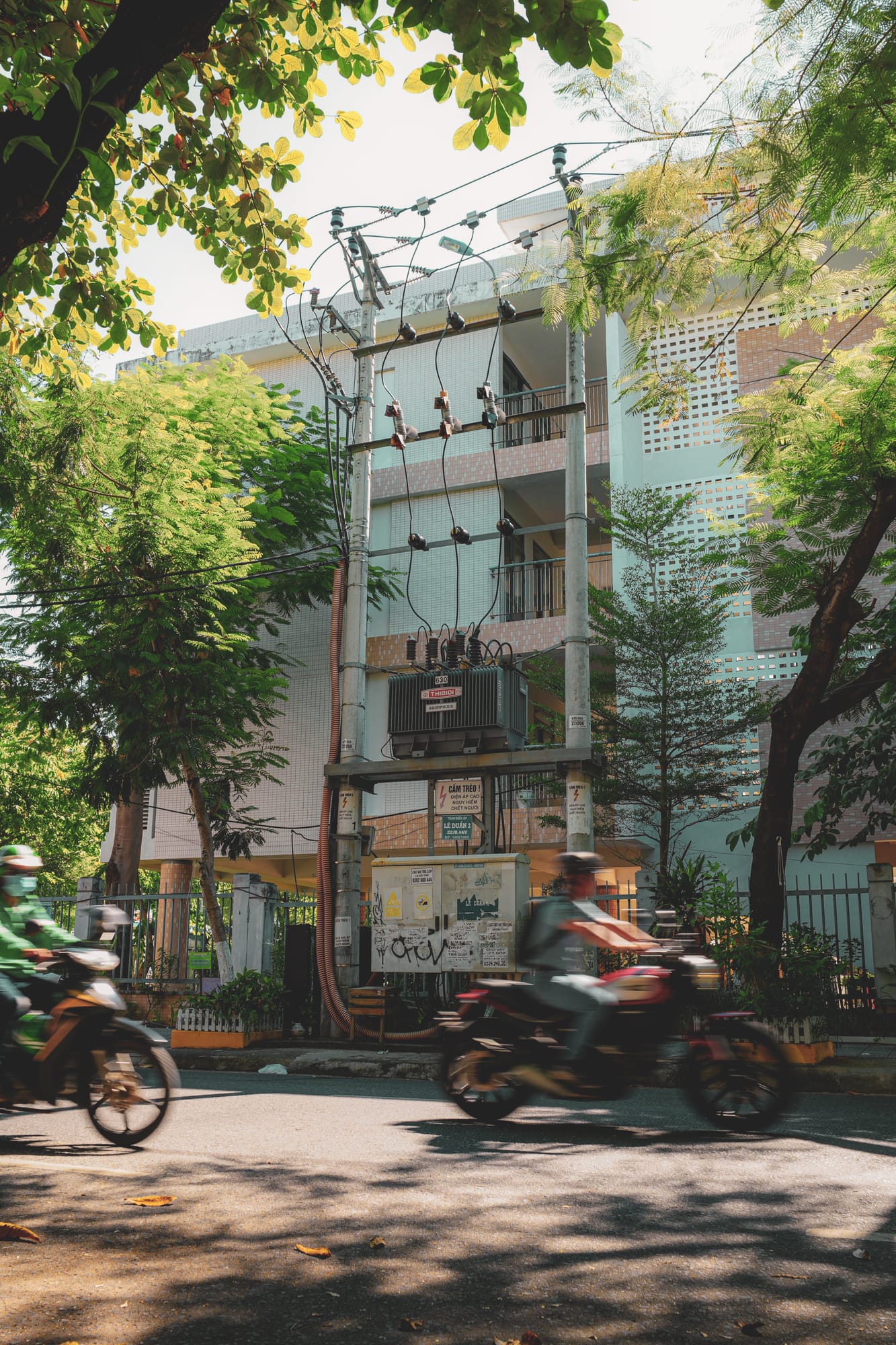 Motorbikes blurring past a mint-green building and a tangle of power lines on a tree-lined street in Vietnam