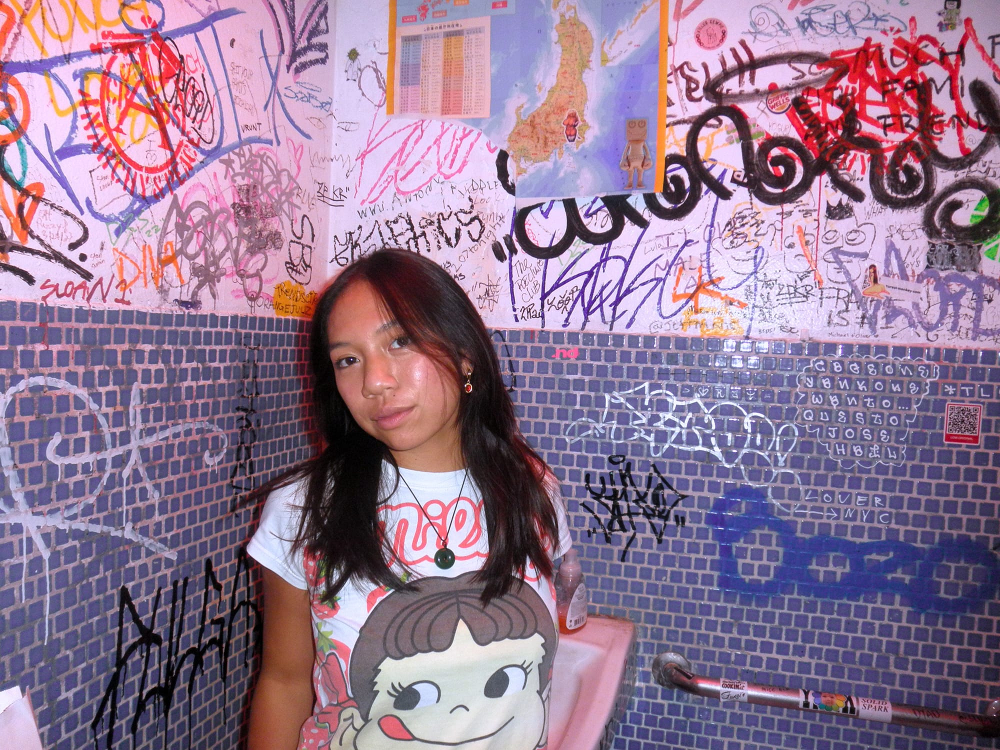 A young woman leaning against a bathroom wall thick with marker graffiti above purple mosaic tile, wearing a white printed tee and a jade pendant