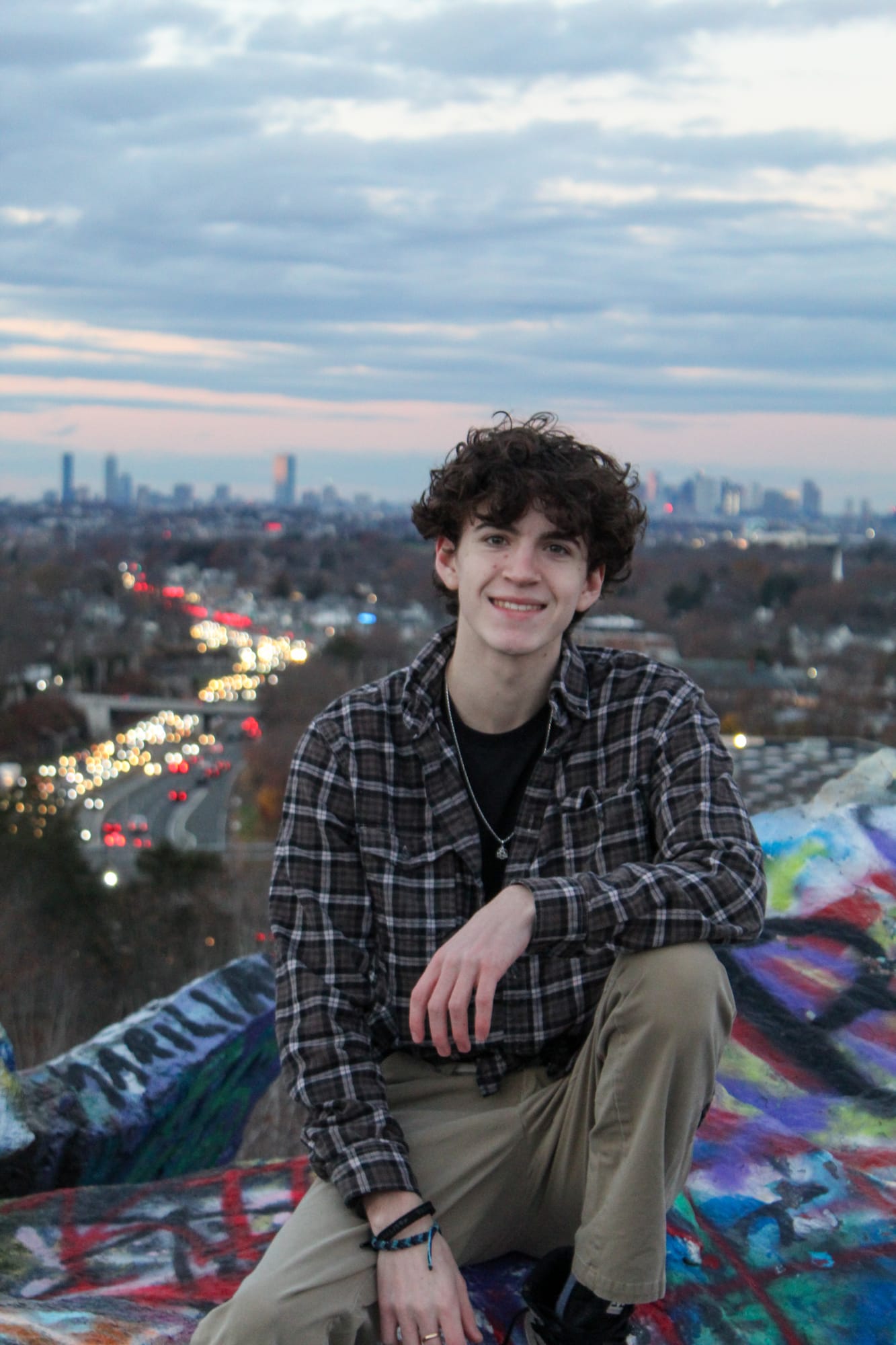 A person in a plaid shirt sitting on a graffiti-painted rock at dusk, with the Boston skyline and a river of headlights behind them