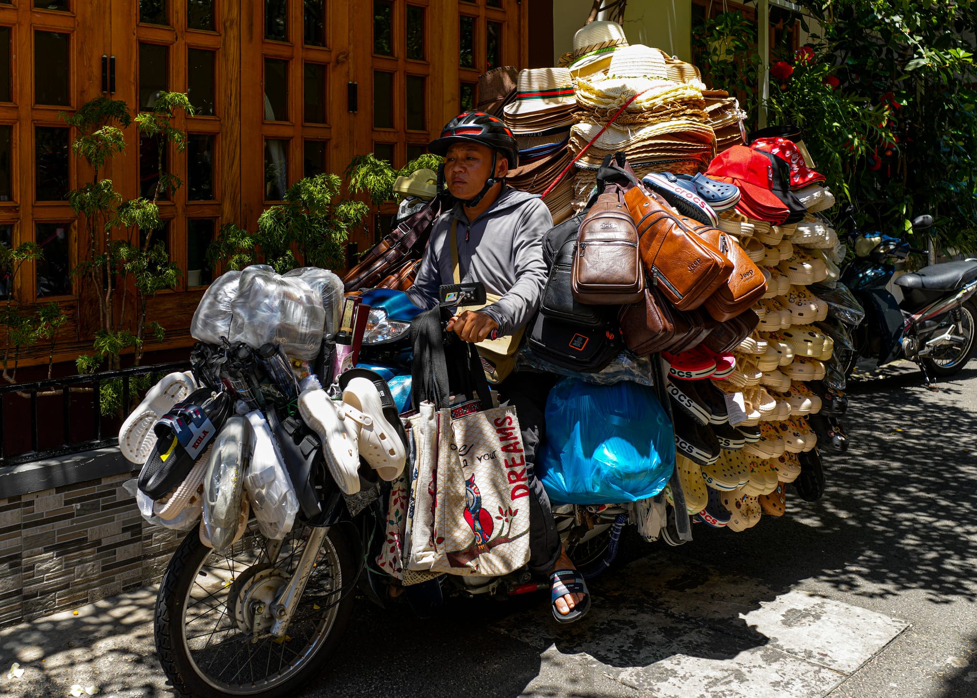 A street vendor on a motorbike loaded shoulder-high with woven sun hats, leather bags and stacks of sandals